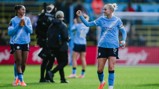 Manchester City Women's team players celebrate on the football field, wearing light blue kits with sponsor logos.