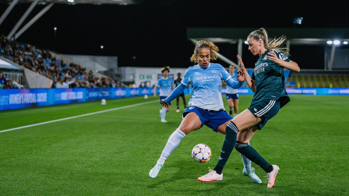 Two soccer players in a match, one from Manchester City Women's team in light blue and one from the opposing team in dark green, compete for the ball near the sideline.