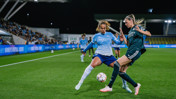 Two soccer players in a match, one from Manchester City Women's team in light blue and one from the opposing team in dark green, compete for the ball near the sideline.