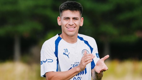 A male football player in a Manchester City jersey clapping with a smile outdoors.