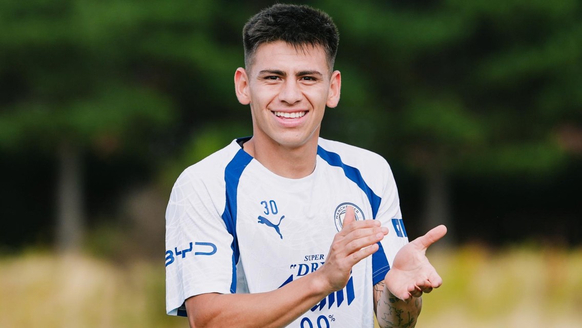 A male football player in a Manchester City jersey clapping with a smile outdoors.