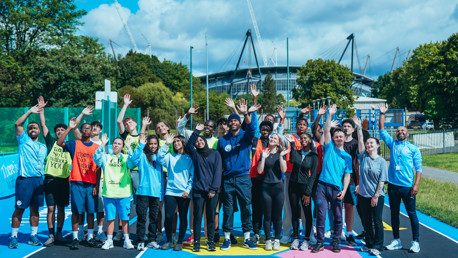 A group of young people with blurred faces standing on a colorful track with their hands raised, near a large stadium in the background.