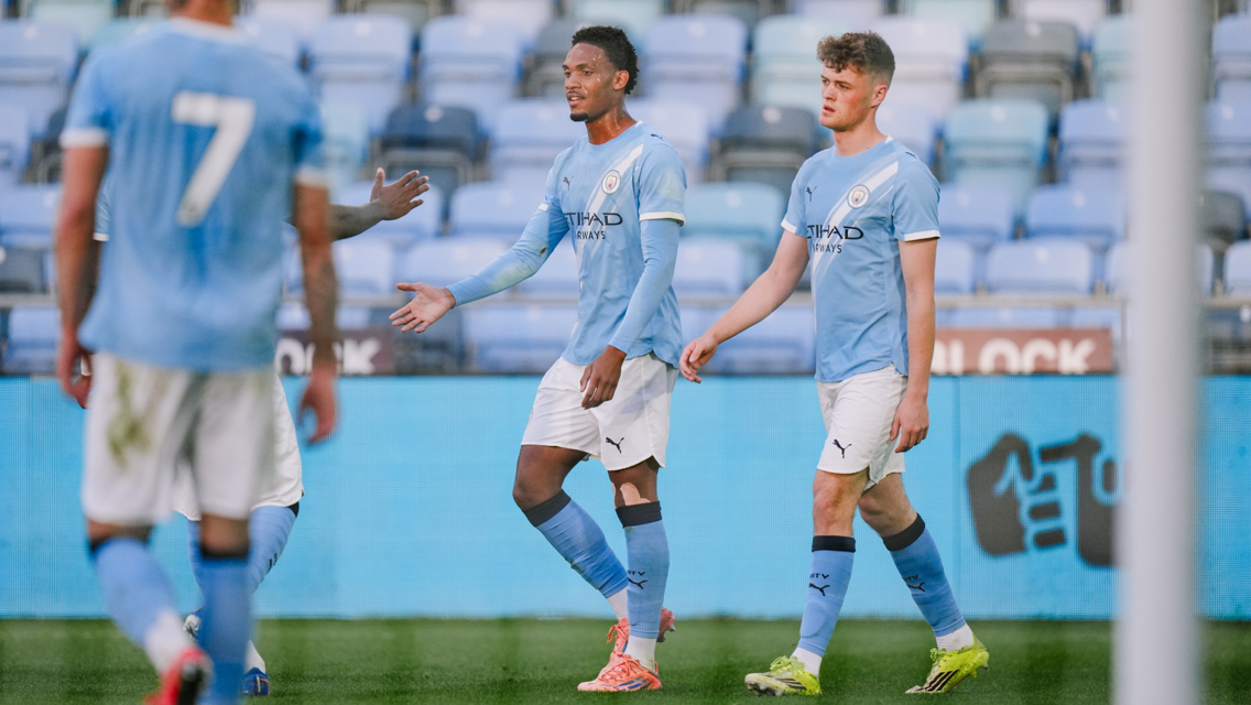 Manchester City players wearing sky blue jerseys during a football match, standing on a pitch in a stadium, one player reaching out for a handshake.