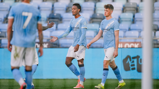 Manchester City players wearing sky blue jerseys during a football match, standing on a pitch in a stadium, one player reaching out for a handshake.