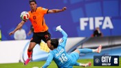 A soccer player from the Netherlands national team wearing jersey number 14 attempts to score against an opposing goalkeeper wearing jersey number 16 at the FIFA City World Cup.