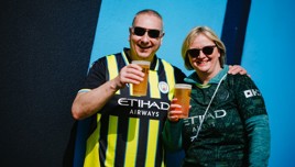 Two people wearing Manchester City jerseys, both holding cups of beer, and smiling against a blue wall.