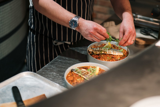 A chef garnishes two dishes with herbs, featuring grilled fish on a bed of a chunky and spiced tomato-based sauce. The chef wears a striped apron in a professional kitchen setting.