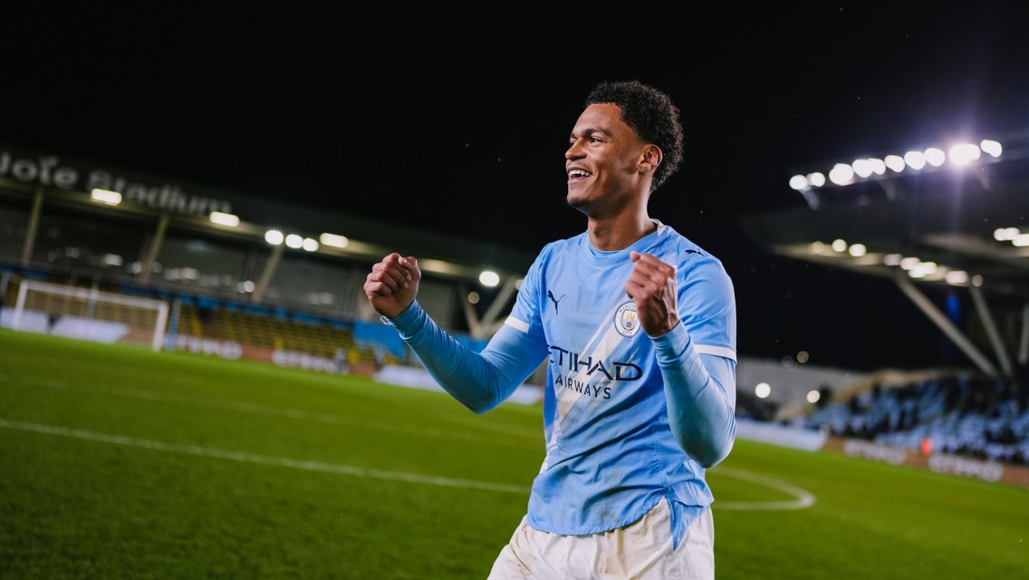 A football player celebrating on the field at night wearing a Manchester City jersey with clenched fists.