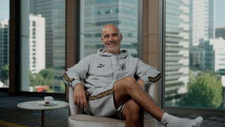 A person smiling while sitting in a chair wearing a grey Puma tracksuit with 'King' on the chest. A window in the background shows a cityscape with tall buildings.