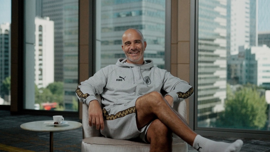 A person smiling while sitting in a chair wearing a grey Puma tracksuit with 'King' on the chest. A window in the background shows a cityscape with tall buildings.