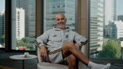 A person smiling while sitting in a chair wearing a grey Puma tracksuit with 'King' on the chest. A window in the background shows a cityscape with tall buildings.