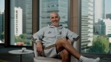 A person smiling while sitting in a chair wearing a grey Puma tracksuit with 'King' on the chest. A window in the background shows a cityscape with tall buildings.