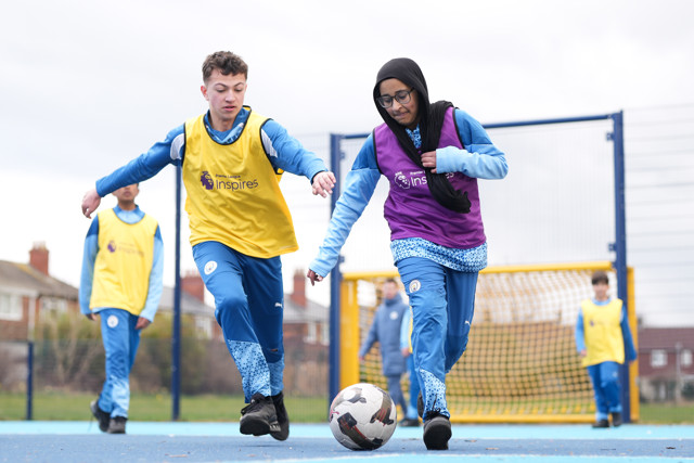 Young players participating in a soccer game wearing Premier League Inspires jerseys in purple and yellow bibs, playing on a colorful outdoor field with netting in the background.