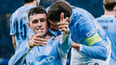 Two Manchester City players in blue jerseys celebrating a goal on the field with arms around each other.