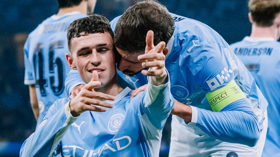 Two Manchester City players in blue jerseys celebrating a goal on the field with arms around each other.
