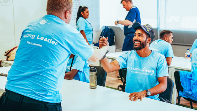 A group of people in light blue t-shirts attending a 'Cityzens Giving Young Leaders Summit' event. Two participants are exchanging a handshake over a table while others are engaged in activities.