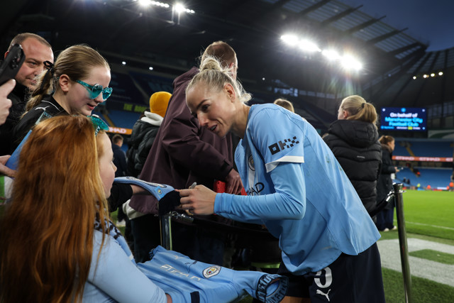 A Manchester City player signing autographs for fans at the Etihad Stadium, engaging with young supporters.