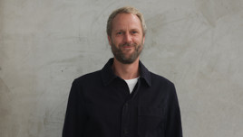 A person wearing a black jacket over a white shirt stands against a concrete wall.