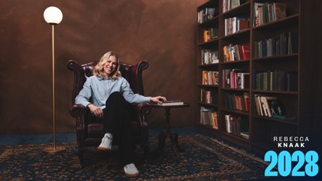 A woman is sitting in a large armchair in a room with a bookshelf filled with books and a floor lamp. The text 'Rebecca Knaak 2028' is in the lower right corner.