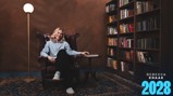 A woman is sitting in a large armchair in a room with a bookshelf filled with books and a floor lamp. The text 'Rebecca Knaak 2028' is in the lower right corner.
