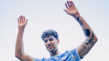 A person wearing a light blue soccer jersey raises their arms, displaying tattoos on both arms.