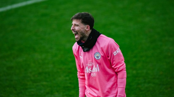 A Manchester City player in a pink training jacket with club logo at a training session on grass field.