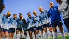 A group of Manchester City women's team players in a huddle wearing light blue uniforms with 'Etihad Airways' logo. The image is taken from a low angle against a clear blue sky.