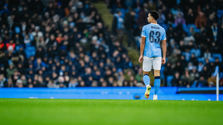 A football player wearing a Manchester City jersey with number 63 and name 'Mukasa' is walking on the pitch. The stadium is filled with spectators watching the match.