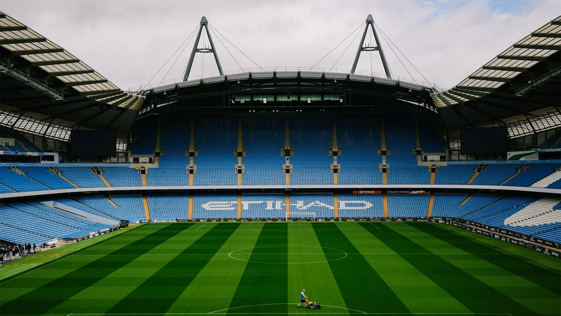 A wide-angle view of the Etihad Stadium showcasing the football pitch, empty blue seats, and stadium roofing structures.