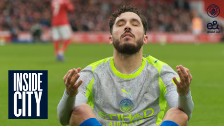 A Manchester City player on the field, wearing a gray and neon green training vest, gestures with his hands during a match. Background shows blurred players and crowd. Includes 'Inside City' text and logos.