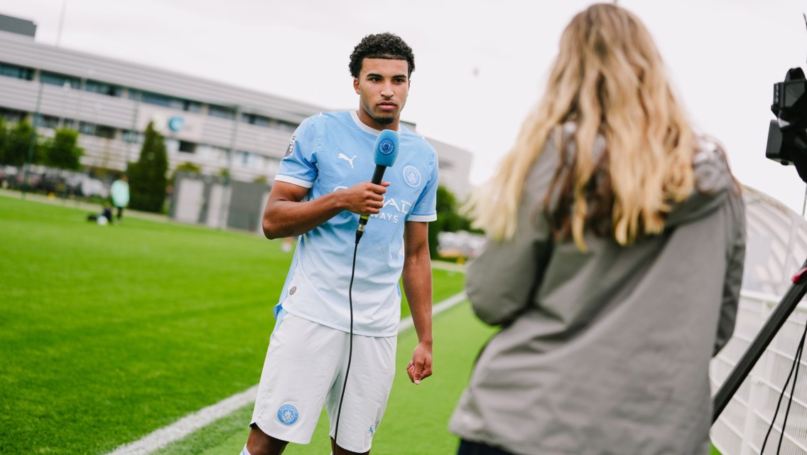 A football player in Manchester City kit is being interviewed on a sports field, holding a microphone. A woman with long hair is facing him, filming with a camera.