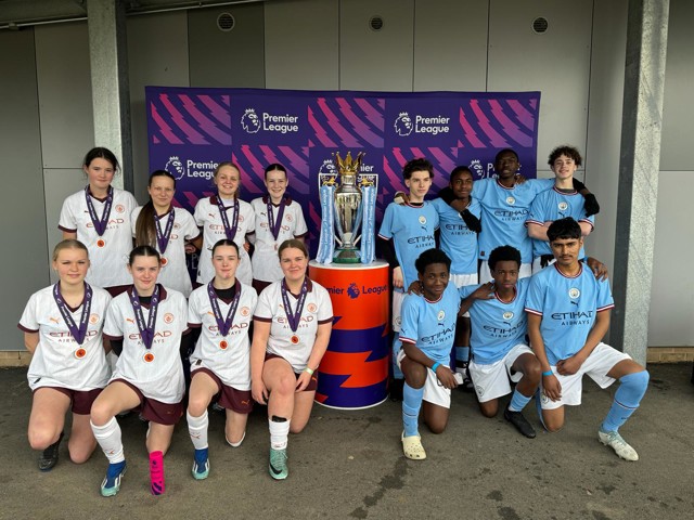 A group photo featuring a youth football team wearing Manchester City and Manchester City Women kits with medals, standing beside the Premier League trophy.