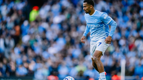 A football player in a Manchester City kit controls the ball on the field during a match, with a blurred crowd in the background.