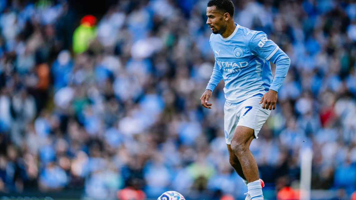 A football player in a Manchester City kit controls the ball on the field during a match, with a blurred crowd in the background.