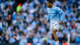 A football player in a Manchester City kit controls the ball on the field during a match, with a blurred crowd in the background.
