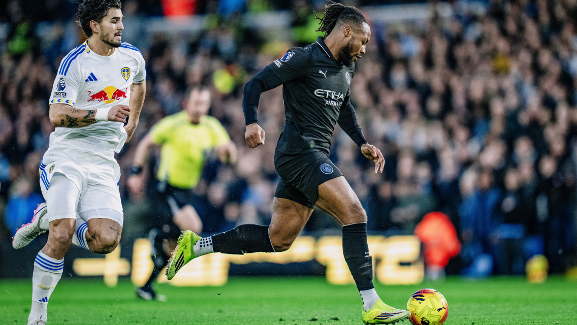 A Manchester City player dribbles the ball pursued by a Leeds United player during a soccer match.