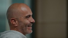 A bald man with a beard wearing a grey hoodie is smiling and looking to the side in a profile view. The background is blurred.
