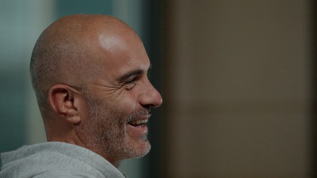 A bald man with a beard wearing a grey hoodie is smiling and looking to the side in a profile view. The background is blurred.