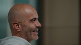 A bald man with a beard wearing a grey hoodie is smiling and looking to the side in a profile view. The background is blurred.