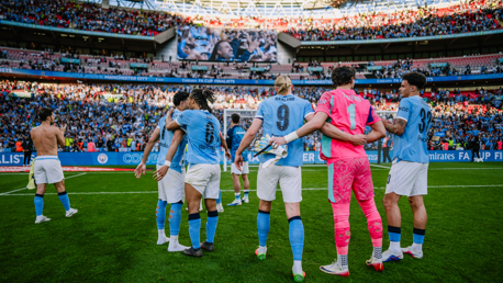 Manchester City players wearing light blue jerseys celebrate on the field in front of a cheering crowd, with 'FA Cup Finalists' displayed in the background. Some players are wearing football gloves, and one is topless.
