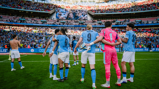 Manchester City players wearing light blue jerseys celebrate on the field in front of a cheering crowd, with 'FA Cup Finalists' displayed in the background. Some players are wearing football gloves, and one is topless.