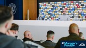 A press conference at Manchester City with a person at the podium, attendees visible in the foreground. The backdrop features multiple sponsor logos.