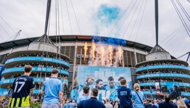 Fans gather in front of the Etihad Stadium, home of Manchester City, during a celebration event, featuring banners, fire effects, and a large crowd.