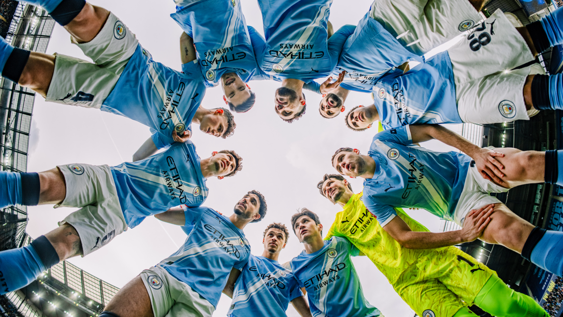 Football players in light blue and white kits stand in a circle, looking downward into the center, likely for a team huddle. The environment suggests they are on a football field.