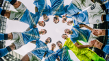 Football players in light blue and white kits stand in a circle, looking downward into the center, likely for a team huddle. The environment suggests they are on a football field.