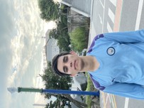A person wearing a light blue Manchester City football jersey with a blurred face, standing on a city street. Background includes trees, buildings, and a cloudy sky.