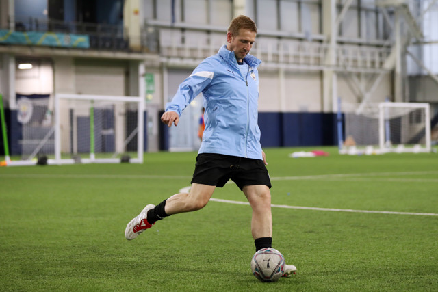 A football player in a blue jacket and black shorts is mid-action kicking a soccer ball in an indoor training facility. The goal and other training equipment are visible in the background.