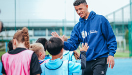 A man wearing a 'City in the Community' hoodie gives a high-five to a group of children on a sports field.
