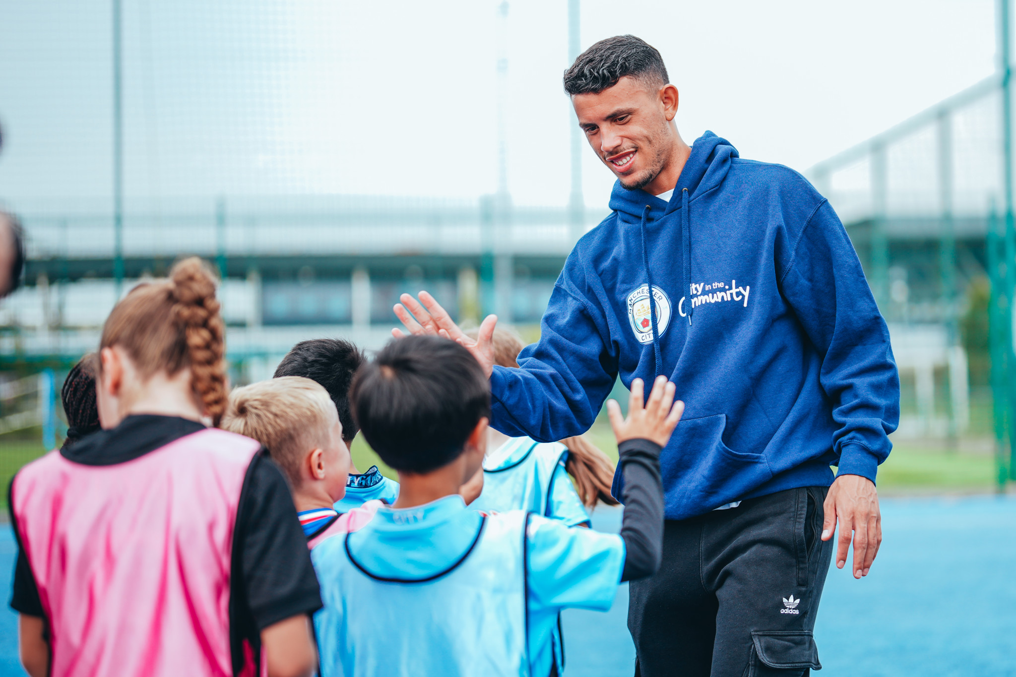 A man wearing a 'City in the Community' hoodie gives a high-five to a group of children on a sports field.