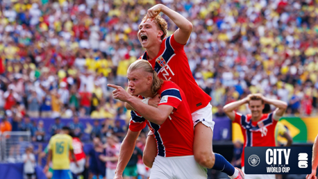 Two football players in red Norway jerseys celebrate a goal, with one player piggybacking on the other. A crowd and other players in similar jerseys are blurred in the background. City World Cup logo is visible.